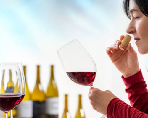 Close up of female enologist smelling wine cork at tasting. Woman holding red wine glass with out of focus bottles in background.